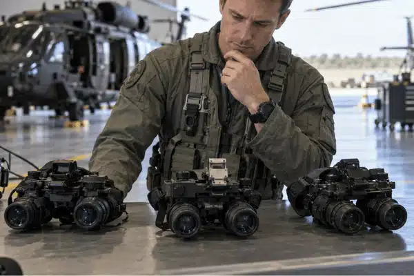 A military aviator examines three night vision goggle systems, including an ANVIS 10 setup, on a workbench inside an aircraft hangar