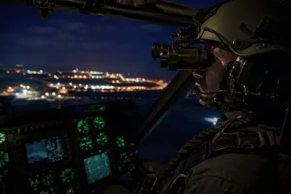 A pilot wearing white phosphor night vision goggles flies a helicopter over city lights at night