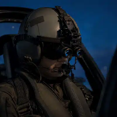 A seated pilot uses helmet mounted night vision goggles while preparing inside a dim aircraft cockpit.