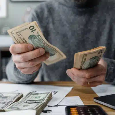 A person counts US dollar bills on a table with paperwork, budget notes, and calculator