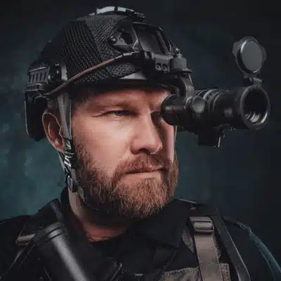 Close-up tactical operator uses helmet-mounted night-vision scope, staring forward in studio portrait wearing protective gear