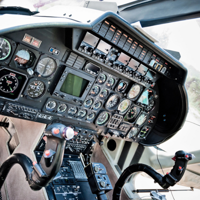 Close-up helicopter cockpit shows analog gauges, switches, and controls used for navigation and flight operations