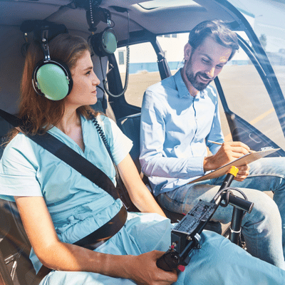 Student pilot practices in helicopter while instructor reviews checklist and cfi syllabus, discussing maneuvers together