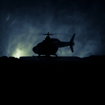 A military helicopter silhouette sits in the dark sky illuminated by dramatic light behind