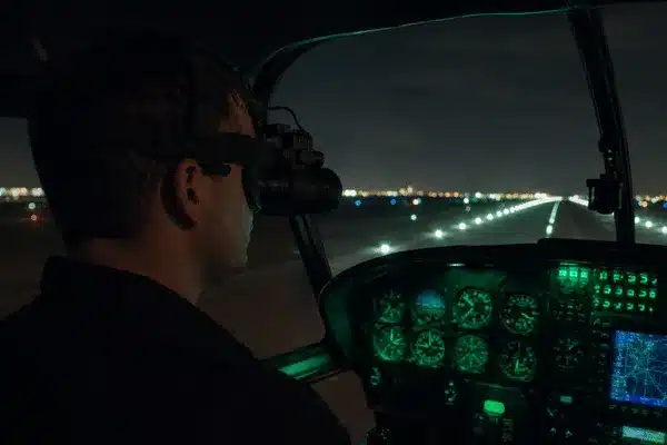 A pilot wearing night vision goggles flies inside a helicopter cockpit during NVG training in Orlando with runway lights ahead