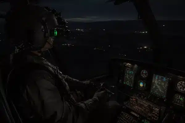 A pilot wearing aviation night vision goggles flies an aircraft over a dark landscape, illustrating how does night vision work during nighttime flight operations.