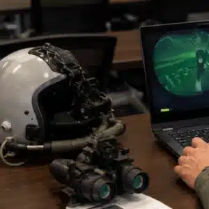 This image shows a pilot using a laptop for night vision goggle online training with a flight helmet and NVGs on the desk
