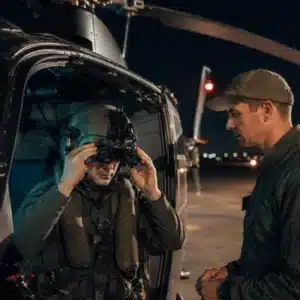 A pilot adjusts night vision goggles beside a helicopter while an instructor observes during NVG training in Orlando