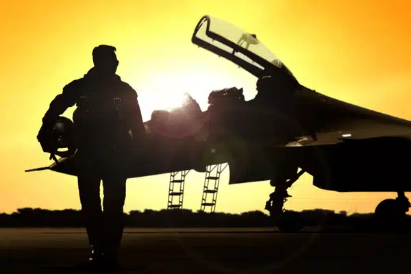 Silhouetted pilot walks toward fighter jet at sunset, carrying helmet and flight goggles beside runway