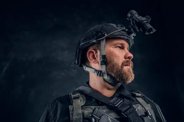 Bearded tactical operator wearing helmet-mounted night-vision device looks left in dramatic studio portrait with gear